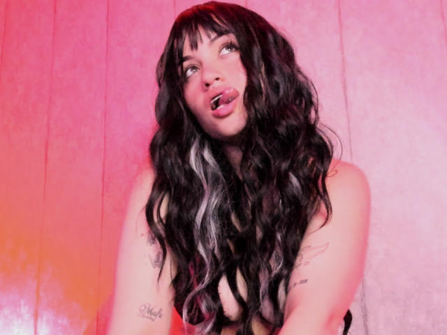 SophieRouge with long, wavy black and white hair, sticking her tongue out, looking up with a playful expression, showing tattoos on her arms, pinkish-red lighting.