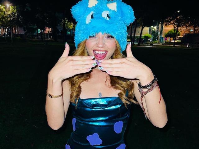 IvyCors with blue fuzzy cat hat, tongue out, hands near face, wearing metallic blue dress with purple polka dots, smiling, showing thumbs up, grassy night setting, colorful bracelets.