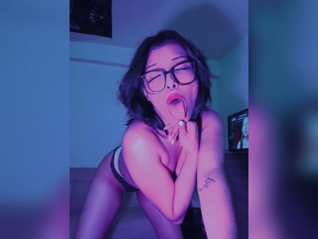 SoySerenaI, with glasses, tongue out, hand near mouth, leaning forward, topless, showing her tits, black underwear, tattoo on left arm, blue-purple light, short dark hair, bedroom.