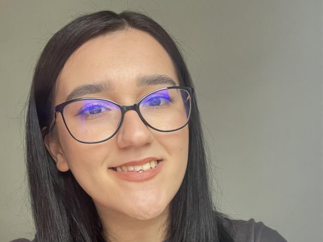 Denise69s has black glasses with blue glowing lenses, straight black hair, and a slight smile, showing a cute face with a light skin tone and a subtle chin dimple.