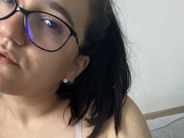 Denise69s with dark hair, glasses, and a slight pout, showing her tits in a white top, against a plain background. Her skin is light, and she's wearing small earrings.