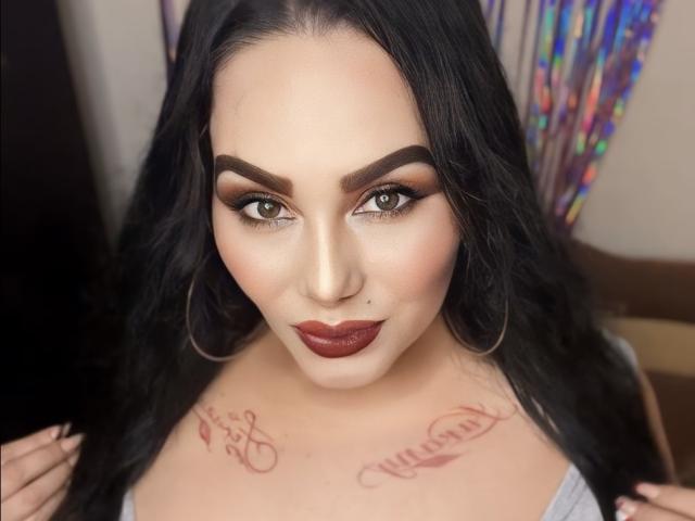 AlondraaHot has dark hair, green eyes, thick eyebrows, and dark red lipstick. She has tattoos on her chest and wears large hoop earrings. Her tits are partially visible.