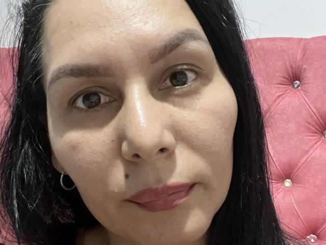 AlondraaHot has a small black beauty mark on her nose, dark brown eyes, and long black hair. She's wearing pink lipstick and small hoop earrings, sitting on a pink tufted chair.