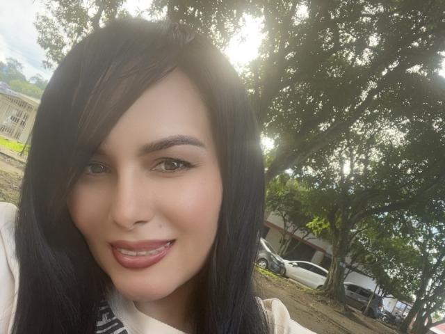 AlondraaHot with black hair, green eyes, and red lipstick, smiling at the camera. Sunlight filters through trees in the background. She has a small nose and full lips.