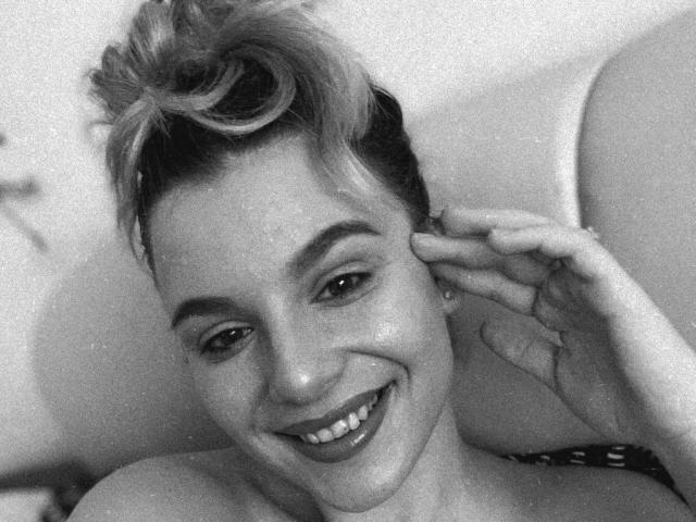 MelanieHartley with short, messy blonde hair, smiling, touching her ear, showing her teeth, and wearing a dark patterned top, in a black-and-white photo.