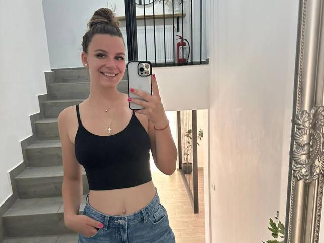 MelanieHartley with her boobs in a black tank top, wearing blue jeans, smiling at a mirror selfie, hand in pocket, pink nails, brown bun, necklace, and white wall.