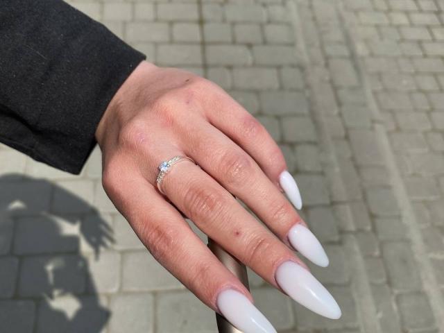 De hand van Kripkoa, met lange, witte, puntige nagels, een zilveren ring op de wijsvinger en een zwarte mouw, op een grijze bakstenen ondergrond..