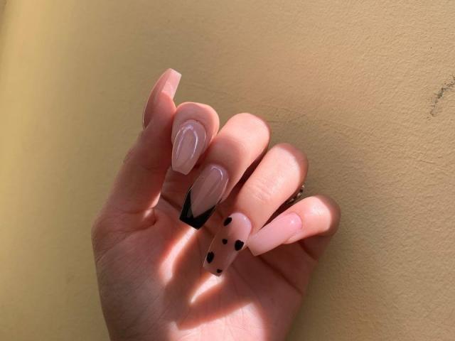 La main de Kripkoa, avec des ongles en forme d'amande, d'un rose pâle et brillant, ornés de pointes noires à motif de points, se trouve contre un mur jaune. La lumière du soleil projette de douces ombres sur ses doigts..
