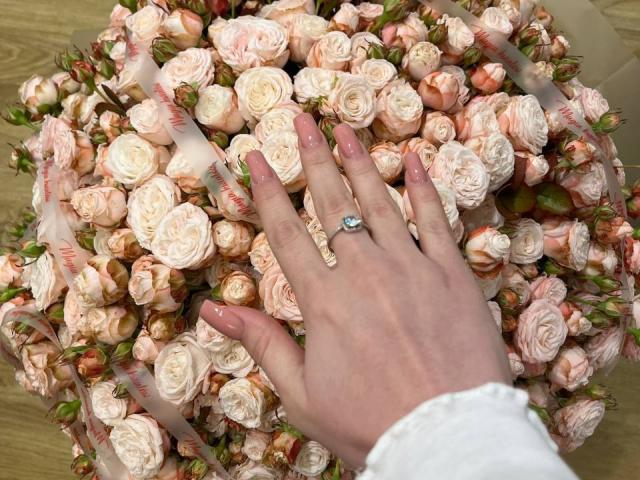 A mão de Kripkoa, com esmalte rosa e um anel de diamantes, toca um buquê de rosas rosa pálido. Seus dedos estão ligeiramente abertos, mostrando a manga branca..