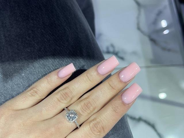 La main de Kripkoa, avec un vernis rose et des ongles carrés, et une bague en diamant sur l'annulaire, repose sur un pantalon gris foncé. Sa peau est claire et ses ongles sont brillants..