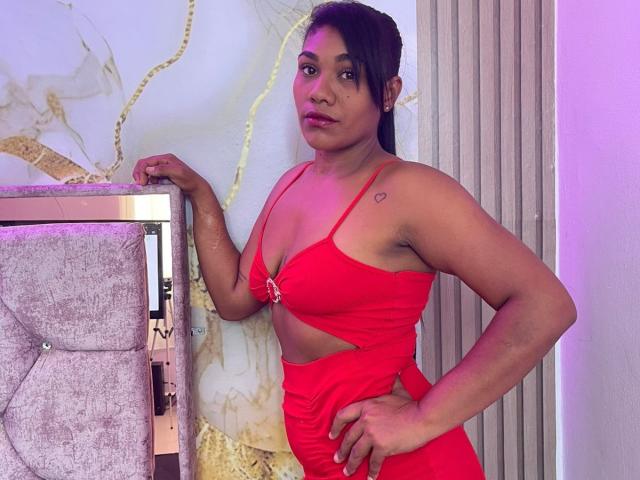 EsmeraldaGarez with medium brown skin and black hair in a ponytail, wearing a red, cut-out dress, showing her tits and a small heart tattoo on her right shoulder.