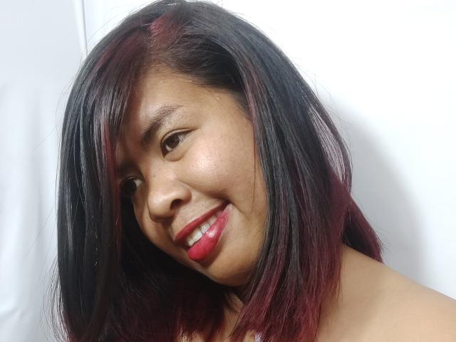 MariahSquirting69 with dark black and red-streaked hair, smiling with red lipstick, brown eyes, and light skin, looking to the side, with a slight tilt of her head.