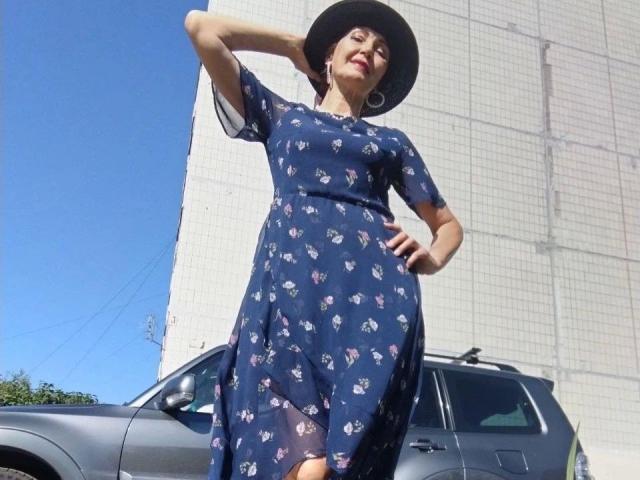 SilviaDP-hot in a blue floral dress, black hat, and hoop earrings, standing by a gray car, hand on hip, looking down confidently with a slight smile.