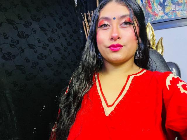 KalpanaAisha with long black hair, red makeup, and red dress, showing off her tits in a selfie. She's got pink lipstick and a bindi on her forehead.