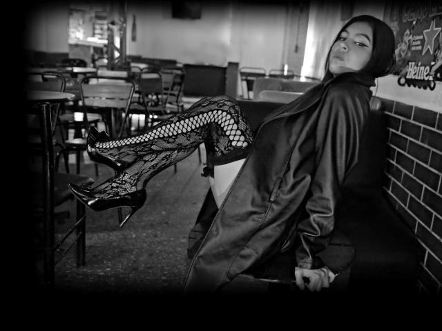 NenaBlack est assise, penchée en arrière dans une veste en cuir, portant des bas résille à motifs de dentelle et des talons hauts noirs, les jambes relevées et avec une expression séduisante. Noir et blanc..