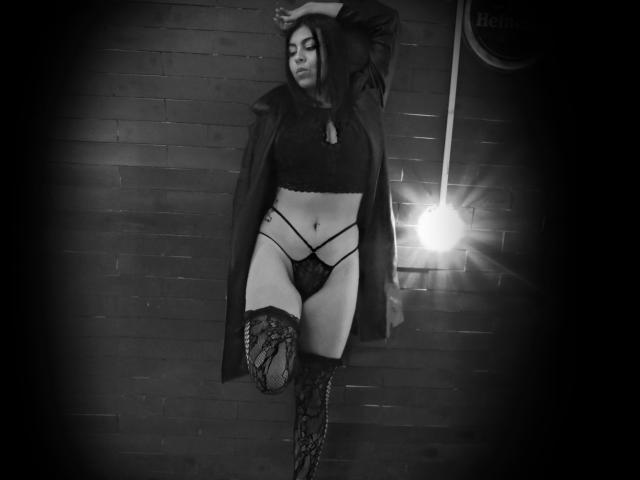 NenaBlack with dark hair, black lace bra, strappy black panties, lace thigh-highs, and open coat, standing, one leg raised, arm raised, in black-and-white, with a light flare.