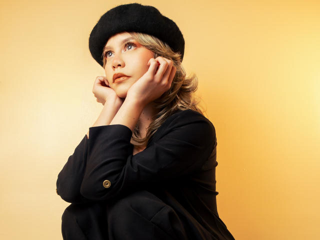 TessaNoir, avec des cheveux blonds ondulés, une casquette noire et une veste noire, les mains sur les joues, fond jaune, regardant vers le haut avec une expression douce, mignonne, élégante, féminine..