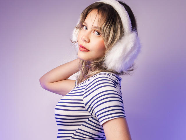 TessaNoir con i capelli biondi, un cappello bianco e soffice a forma di orecchie, e una camicia a righe, che si guarda indietro, mostrando il sedere, con un leggero sorriso, su uno sfondo viola sfumato..