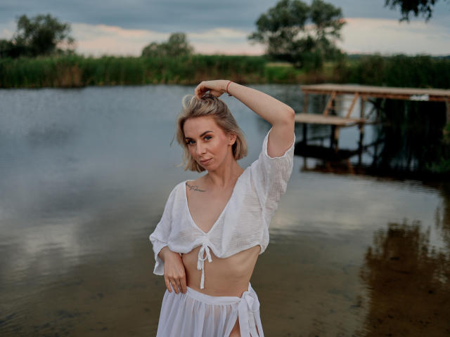 VasilisaSexy with blonde hair, white sheer top, and white skirt, standing by a lake, hand in hair, small tits, tattoo on chest, water reflecting her, wooden dock in background.