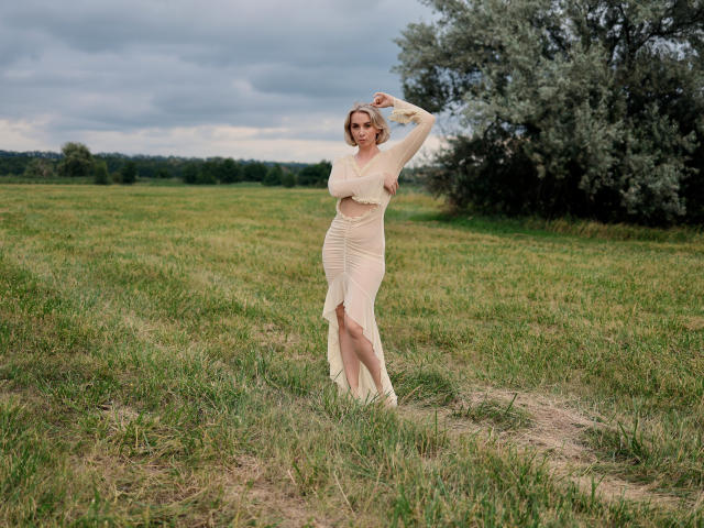 VasilisaSexy, standing in a grassy field, wearing a sheer, beige, long-sleeve dress, with one arm raised, showing her tits and a slight side slit.