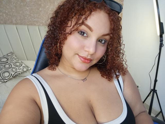 YeraCarey tiene el pelo rojo rizado, aros grandes, una camiseta negra y blanca y un collar. Está sentada con una ligera sonrisa, mostrando sus grandes senos y su lápiz labial rosa..