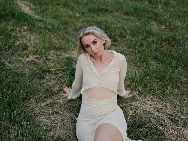 VasilisaSexy with blonde hair, wearing a sheer, white, cutout dress, sitting on grass, arms behind her, looking at camera, slightly smiling, green grass background.
