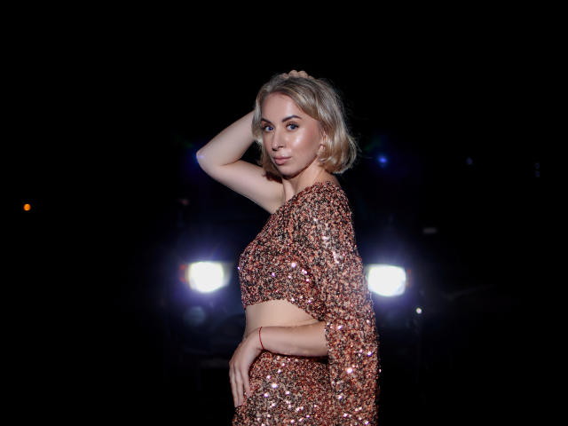 VasilisaSexy with blonde hair, wearing a sparkly, sequined, one-shoulder, midriff-baring dress, hand in hair, standing in dark with two bright lights in background.