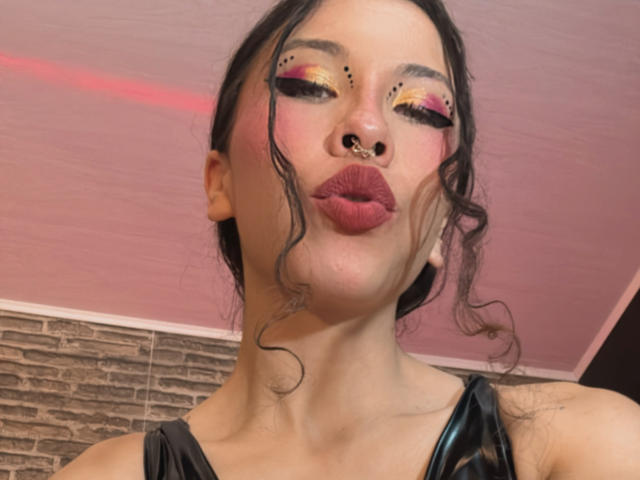 SofiBrandford has dark wavy hair, bold pink and yellow eyeshadow, black eyeliner, a nose ring, and puffy red lips, wearing a black leather top, kissing the camera.