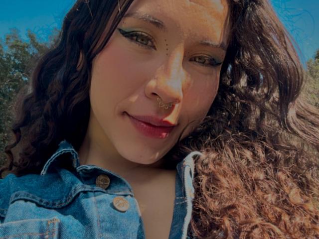 SofiBrandford with curly brown hair, wearing a blue denim jacket, has a nose ring and black winged eyeliner, smiling slightly, under bright sunlight.