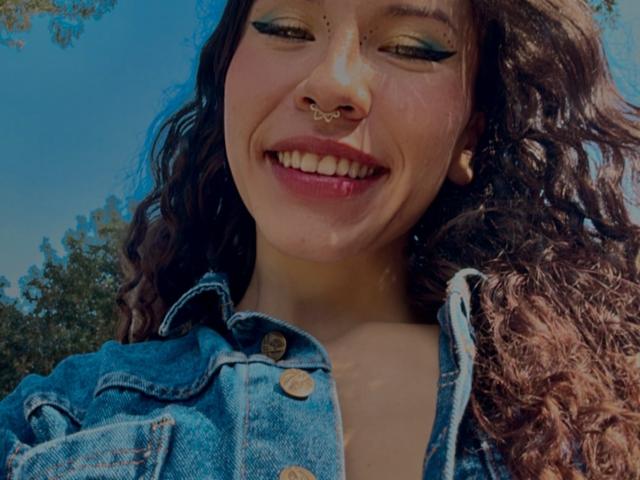 SofiBrandford with curly brown hair, blue denim jacket, green eyeshadow, nose ring, smiling, showing teeth, and red lipstick, looking down at camera.