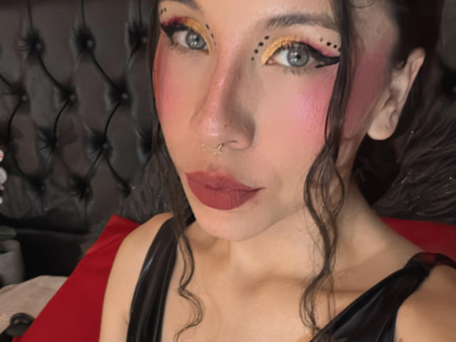 SofiBrandford has pink-tinted skin, black eyeliner with yellow accents, dark red lipstick, and wavy brown hair. She's wearing a black top and has a nose ring.