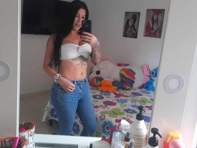 SexyCaandydoll with long black hair, white strapless top, showing her tits, tattoo on right side, blue jeans, standing in mirror, holding phone, smiling, bedroom with colorful bedspread.