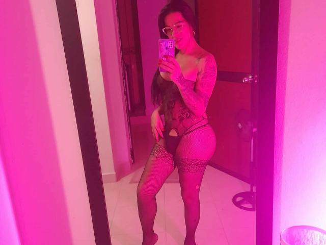 SexyCaandydoll with tattoos, wearing lace lingerie, leopard-print thigh-highs, and glasses, taking a mirror selfie, showing her ass and small boobs, in pink-lit room.