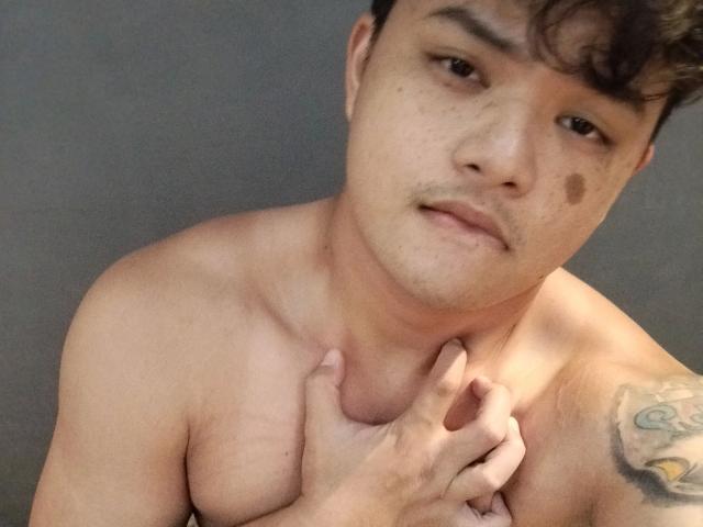 Shirtless AsianFuckboy-hot with curly brown hair, medium skin, small mole on cheek, tattoo on right shoulder, holding left chest with right hand, looking slightly bored.