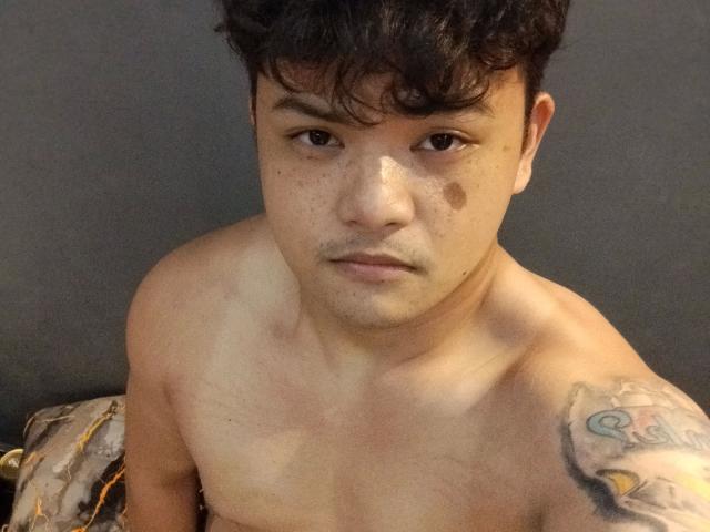 AsianFuckboy-hot with short, curly black hair, freckled face, small brownish birthmark on left cheek, tattoo on right shoulder, and bare chest, looking straight into camera.