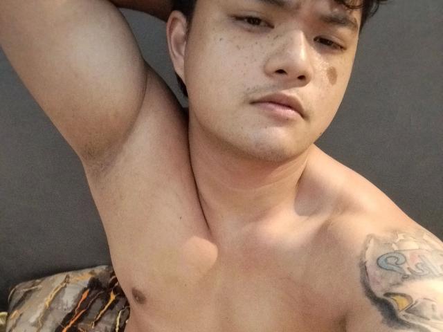AsianFuckboy-hot, shirtless with arms raised, showing a tattoo on his right shoulder, has a slightly chubby face, brown eyes, and short curly black hair. His armpits are hairy.