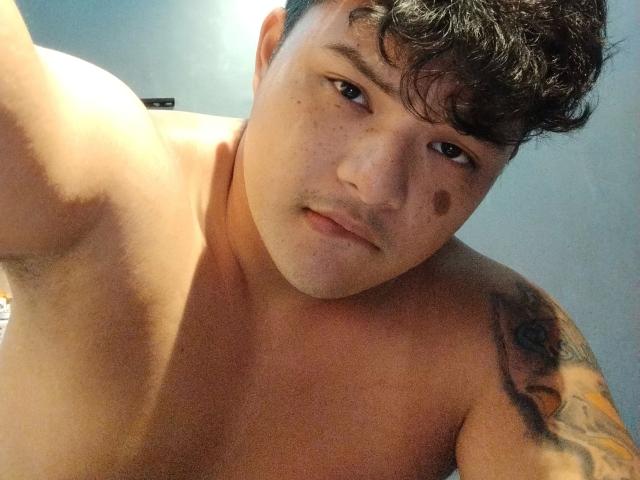 AsianFuckboy-hot with short, curly black hair, freckles, and a tattoo on his right shoulder, looking down at the camera with a slightly confused expression, shirtless.
