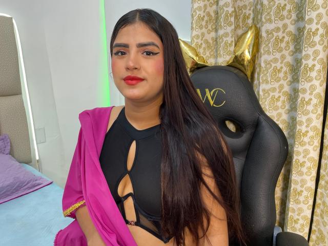 NayaYalitza with long dark hair, red lipstick, and black cut-out top, pink shawl, sitting in black chair with gold star, looking at camera, bed and curtains in background.