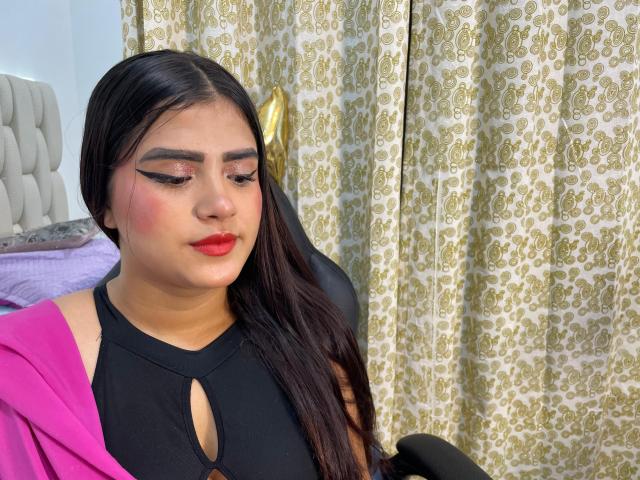 NayaYalitza with long black hair, red lipstick, and black cut-out top, wearing pink jacket, sitting in chair, looking down, patterned curtains in background, subtle eye makeup, soft skin.