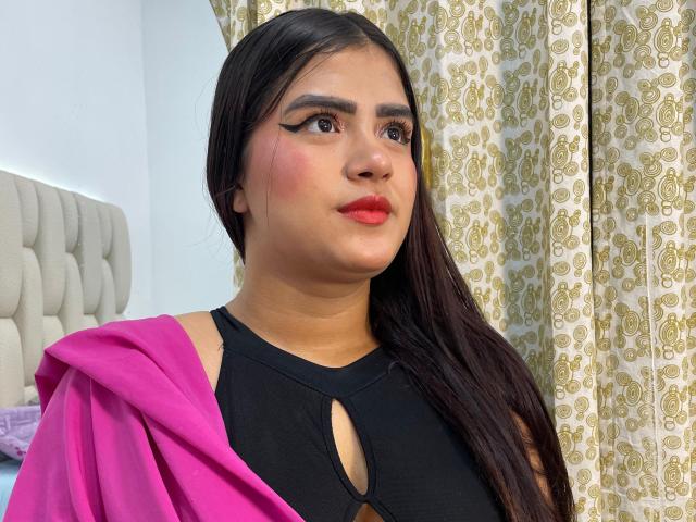 NayaYalitza with long dark hair, red lipstick, black cut-out top, pink jacket, and winged eyeliner, looking up with a slightly pouty expression. Her tits are small, visible through top.