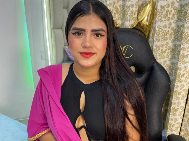 NayaYalitza with long black hair, red lipstick, and black cutout top, wearing pink shawl, sitting in black gaming chair, looking at camera, slightly smiling.