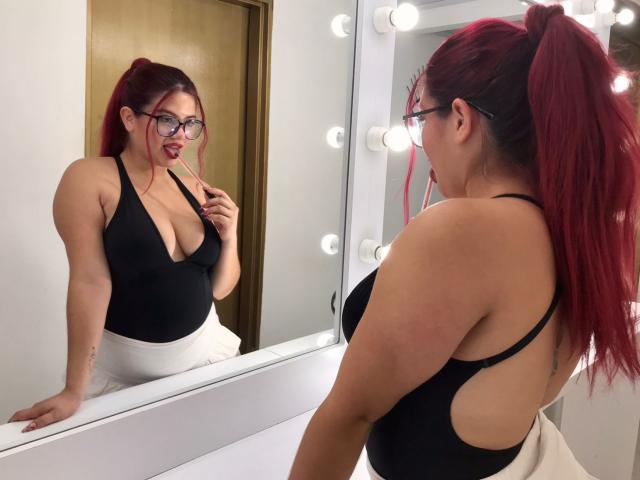 RubyMartin with red hair in a ponytail, wearing glasses and a black low-cut top, showing her tits in the mirror, touching her red lipstick.