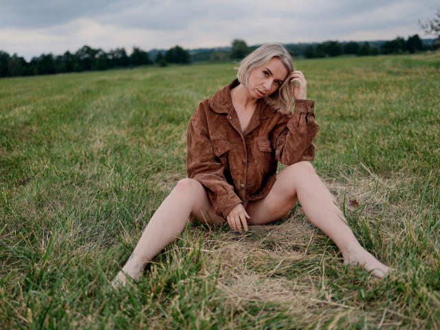 VasilisaSexy with blonde hair, brown shirt, and no pants, sitting on grass with legs spread, hand in hair, looking at camera, showing her pussy and bare ass.