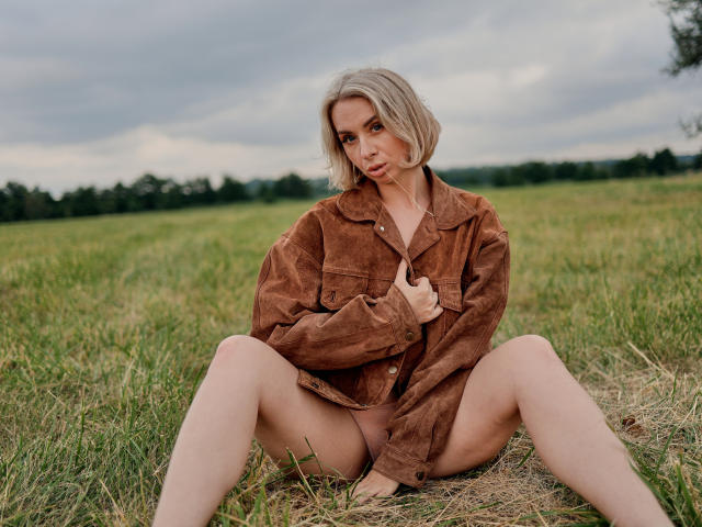 VasilisaSexy sitting in grass with legs spread, wearing a brown suede jacket, blonde bob, and no pants, showing her pussy. Overcast sky, green field in background.