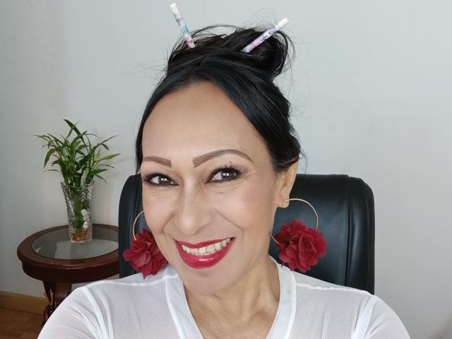 GinaOne has dark hair in a bun with hairpins, red lipstick, and red flower earrings, wearing a white shirt, smiling with visible makeup, and a small plant in the background.