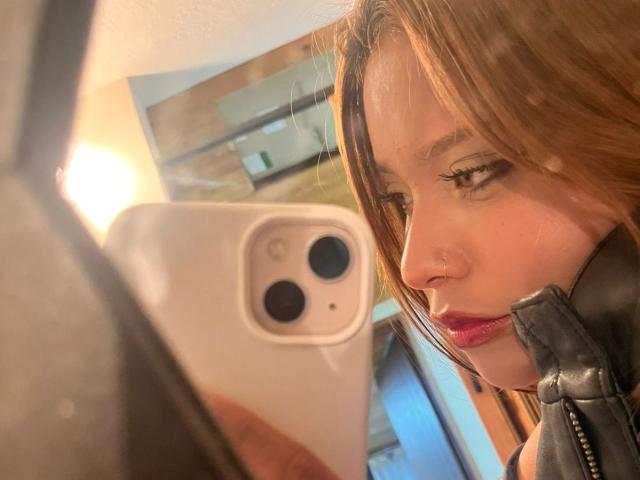 HeraJuno with light brown hair, red lipstick, and black leather jacket, taking a selfie in a mirror with an iPhone, her nose ring visible, her expression slightly pouty.