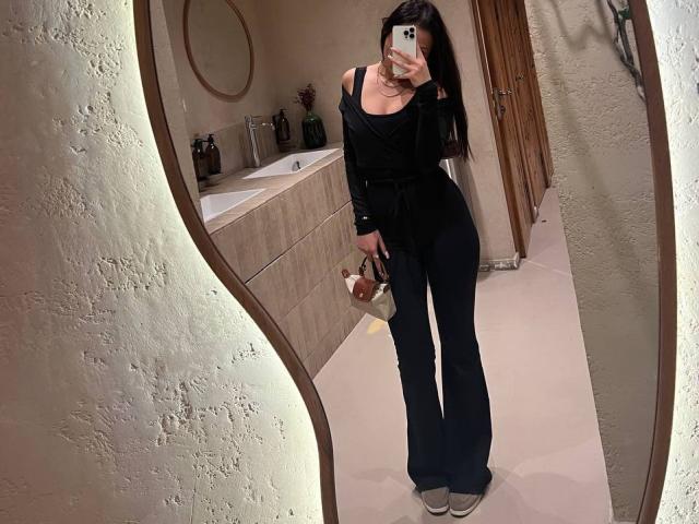 NaomiAsian with long black hair, off-shoulder black top, and flared black pants, holding a purse, taking a mirror selfie in a bathroom, showing her ass and small tits.