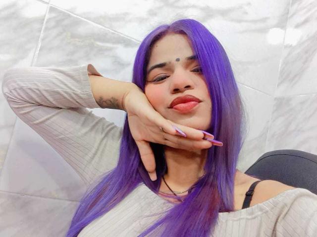 KairaDayamai with purple hair, pouty lips, and a tattoo on her left hand, wearing a beige ribbed top, resting her chin on her hand. Boobs partially visible.