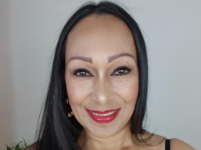 GinaOne has dark brown eyes, thick black eyebrows, red lipstick, and black hair with a small mole above her right eye. She's smiling with her tits barely visible.