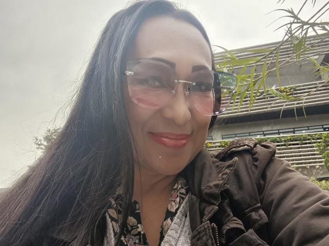 GinaOne has long black hair, pink-tinted glasses, red lipstick, and a slight smile. She wears a dark jacket over a floral shirt. Her nose is slightly shiny.